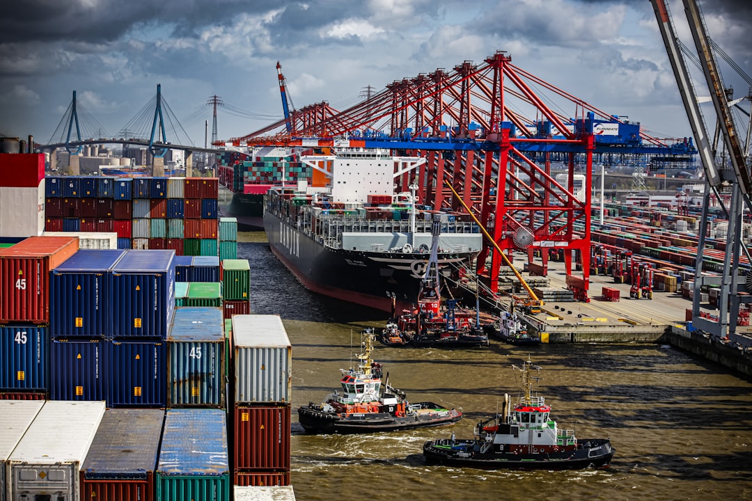 a tug boat in the water next to a large cargo ship, Vibrant port scene with a large cargo ship being guided by tugboats, flanked by towering red cranes and stacks of multicolored shipping containers under a dynamic sky.