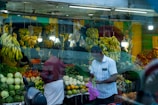 A busy fruit and vegetable market stall with bananas hanging prominently above a variety of produce including cabbages, tomatoes, and other greens. Two men are present; one is holding a plastic bag with some vegetables, while the other has his back turned. Dim lights illuminate the space, and a scooter is parked in front.