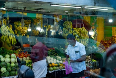 A busy fruit and vegetable market stall with bananas hanging prominently above a variety of produce including cabbages, tomatoes, and other greens. Two men are present; one is holding a plastic bag with some vegetables, while the other has his back turned. Dim lights illuminate the space, and a scooter is parked in front.