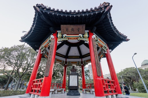 A traditional Chinese pavilion with vibrant red pillars and intricate black roof tiles, situated in a park setting. The structure features detailed decorations and inscriptions under the roof, creating a harmonious blend of cultural architecture and nature. Trees and modern city elements are visible in the background.