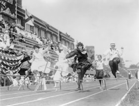 A group of people in historical attire are participating in a hurdle race on a track. The setting appears to be an early 20th-century athletic event. The participants, dressed in various styles including dresses and sailor outfits, are mid-jump over the hurdles. In the background, a crowd is seated in the stands, some under umbrellas.