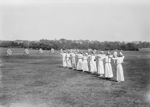 A group of women dressed in light-colored attire stand in a line on a grass field, each holding a bow aimed at multiple archery targets set up at a distance. The attire appears to be from an earlier time period. The setting seems peaceful, with trees and some buildings visible in the background under a clear sky.