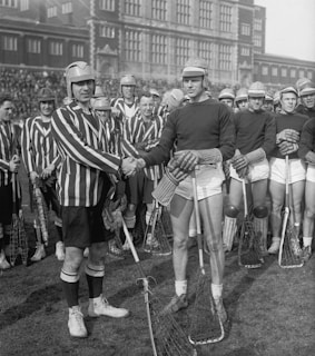 A group of lacrosse players is gathered on a field in front of a large brick building. Two players, one in a striped jersey and the other in a plain dark jersey, are shaking hands in the foreground. Both teams are wearing helmets and holding lacrosse sticks. The players standing behind them appear to be from different teams as well.