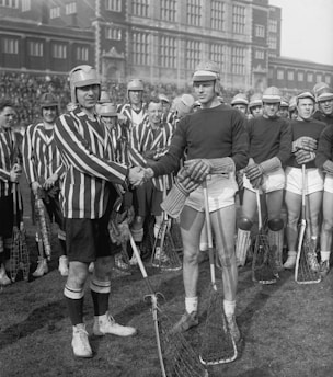 A group of lacrosse players is gathered on a field in front of a large brick building. Two players, one in a striped jersey and the other in a plain dark jersey, are shaking hands in the foreground. Both teams are wearing helmets and holding lacrosse sticks. The players standing behind them appear to be from different teams as well.