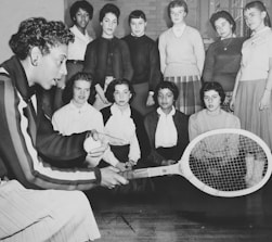 A group of young women gather around a woman who is explaining tennis techniques using a racket and ball. The setting appears educational, with the women attentively watching and listening. The clothing and hairstyles suggest a mid-20th-century style.