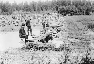 A group of people are engaged in a shooting range activity in an outdoor setting surrounded by trees. Men are lying prone on the ground, aiming rifles, while others stand or sit nearby observing. The scene is captured in a black and white photograph, suggesting a historic or early 20th-century event.