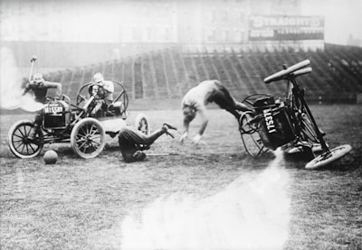 A chaotic scene on a stadium field where a person is being thrown forward from an overturned car labeled 'ALESIL,' with another individual somersaulting mid-air and a third person occupying a different vehicle. A fourth figure is seen behind the second car, seemingly holding a stick. The background shows empty stadium seating.