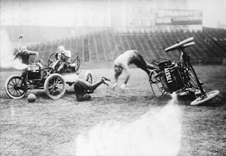 A chaotic scene on a stadium field where a person is being thrown forward from an overturned car labeled 'ALESIL,' with another individual somersaulting mid-air and a third person occupying a different vehicle. A fourth figure is seen behind the second car, seemingly holding a stick. The background shows empty stadium seating.