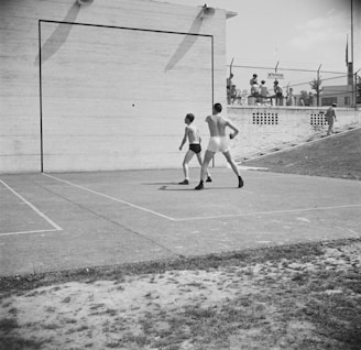 Two men are playing handball on an outdoor court in front of a large, smooth wall. They are dressed in athletic wear, including shorts and sneakers. In the background, several people are sitting and standing near a fence, possibly spectating.