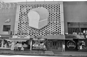 A street scene features a building facade with a large, stylized slice of bread logo, indicating a business named 'Mil Toast House.' Adjacent stores include an eye clinic and a coffee shop. A few people are visible on the sidewalk, some wearing face masks, against a patterned backdrop.