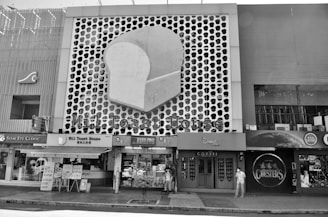 A street scene features a building facade with a large, stylized slice of bread logo, indicating a business named 'Mil Toast House.' Adjacent stores include an eye clinic and a coffee shop. A few people are visible on the sidewalk, some wearing face masks, against a patterned backdrop.