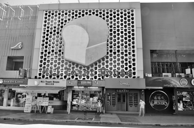 A street scene features a building facade with a large, stylized slice of bread logo, indicating a business named 'Mil Toast House.' Adjacent stores include an eye clinic and a coffee shop. A few people are visible on the sidewalk, some wearing face masks, against a patterned backdrop.