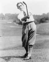 A vintage photograph of a golfer in mid-swing, wearing traditional early 20th-century attire including a white shirt, tie, and knickerbockers. The background features a blurred outdoor scene, likely a golf course.