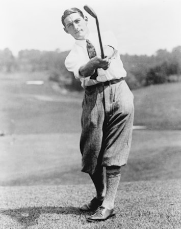 A vintage photograph of a golfer in mid-swing, wearing traditional early 20th-century attire including a white shirt, tie, and knickerbockers. The background features a blurred outdoor scene, likely a golf course.