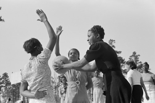 A group of women are actively playing basketball outdoors, with one woman holding the ball while another raises her arms to block. The setting appears to be casual, likely in a practice or friendly game. Trees are visible in the background, suggesting the scene takes place in an open, natural environment.