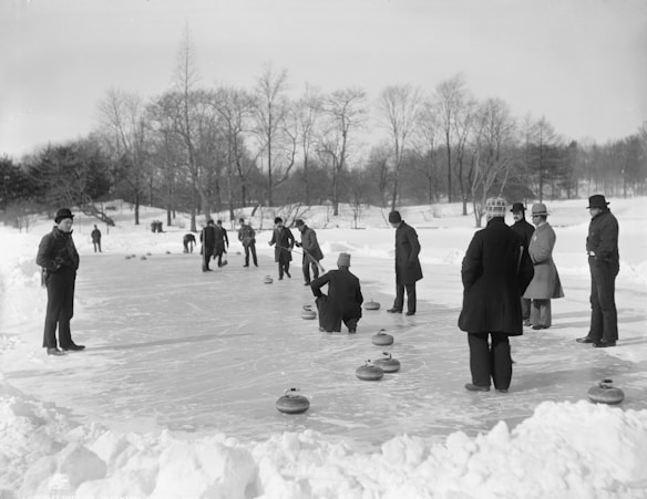 A group of people engaged in a game of curling on an ice-covered surface surrounded by snow. The participants, dressed in winter clothing and hats, are intently focused on the curling stones. The scene is set outdoors in a park-like area with barren trees in the background, reflecting a winter atmosphere.