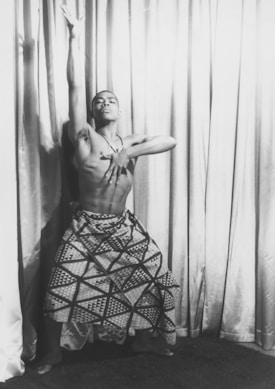 A dancer wearing a patterned skirt strikes a dramatic pose. The dancer's left arm is extended upwards, while the right hand is placed on the chest. The backdrop consists of draped curtains, creating a theatrical atmosphere.