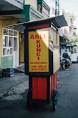 A mobile key duplication cart is situated on a street, featuring a sign with bold red lettering on a bright yellow background. The cart is equipped with wheels, allowing it to be easily moved. The surrounding area includes a sidewalk and buildings with various colors and architectural details. A person is riding a motorcycle in the background.