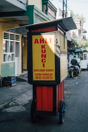 A mobile key duplication cart is situated on a street, featuring a sign with bold red lettering on a bright yellow background. The cart is equipped with wheels, allowing it to be easily moved. The surrounding area includes a sidewalk and buildings with various colors and architectural details. A person is riding a motorcycle in the background.
