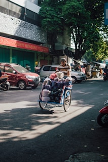 A person wearing a striped shirt pushes a blue cart filled with various colorful bags on a busy street. Several cars and motorcycles are parked along the road, and the background features a shop with a red sign and a tall tree casting shadows.