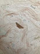 A topographic map featuring contour lines indicating elevation changes, with labels for elevation values. The term 'North' is marked on the map. There are some small blue areas that likely represent water bodies, and a larger brown area possibly representing a peak or significant landscape feature.