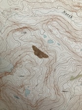 A topographic map featuring contour lines indicating elevation changes, with labels for elevation values. The term 'North' is marked on the map. There are some small blue areas that likely represent water bodies, and a larger brown area possibly representing a peak or significant landscape feature.