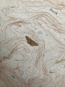 A topographic map featuring contour lines indicating elevation changes, with labels for elevation values. The term 'North' is marked on the map. There are some small blue areas that likely represent water bodies, and a larger brown area possibly representing a peak or significant landscape feature.