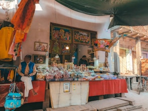 A street vendor's stall filled with a variety of items including snacks, sweets, and spices displayed in transparent bags and containers. Colorful traditional cloths and artwork are hung around the stall, depicting religious or cultural themes. Two people are present, one seated and observing their surroundings, and another attending to the stall behind. A bicycle is leaning against the wall in the right background, and QR payment options are displayed prominently.