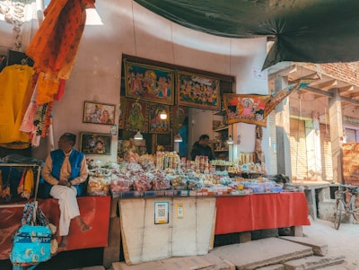 A street vendor's stall filled with a variety of items including snacks, sweets, and spices displayed in transparent bags and containers. Colorful traditional cloths and artwork are hung around the stall, depicting religious or cultural themes. Two people are present, one seated and observing their surroundings, and another attending to the stall behind. A bicycle is leaning against the wall in the right background, and QR payment options are displayed prominently.