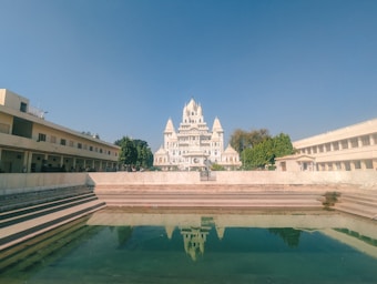 A grand white temple with intricate architecture is situated at the center, flanked by two simple, beige buildings with multiple rooms. In front of the temple, there is a rectangular water body reflecting parts of the temple. Trees add greenery around the temple, and the sky is a clear blue.