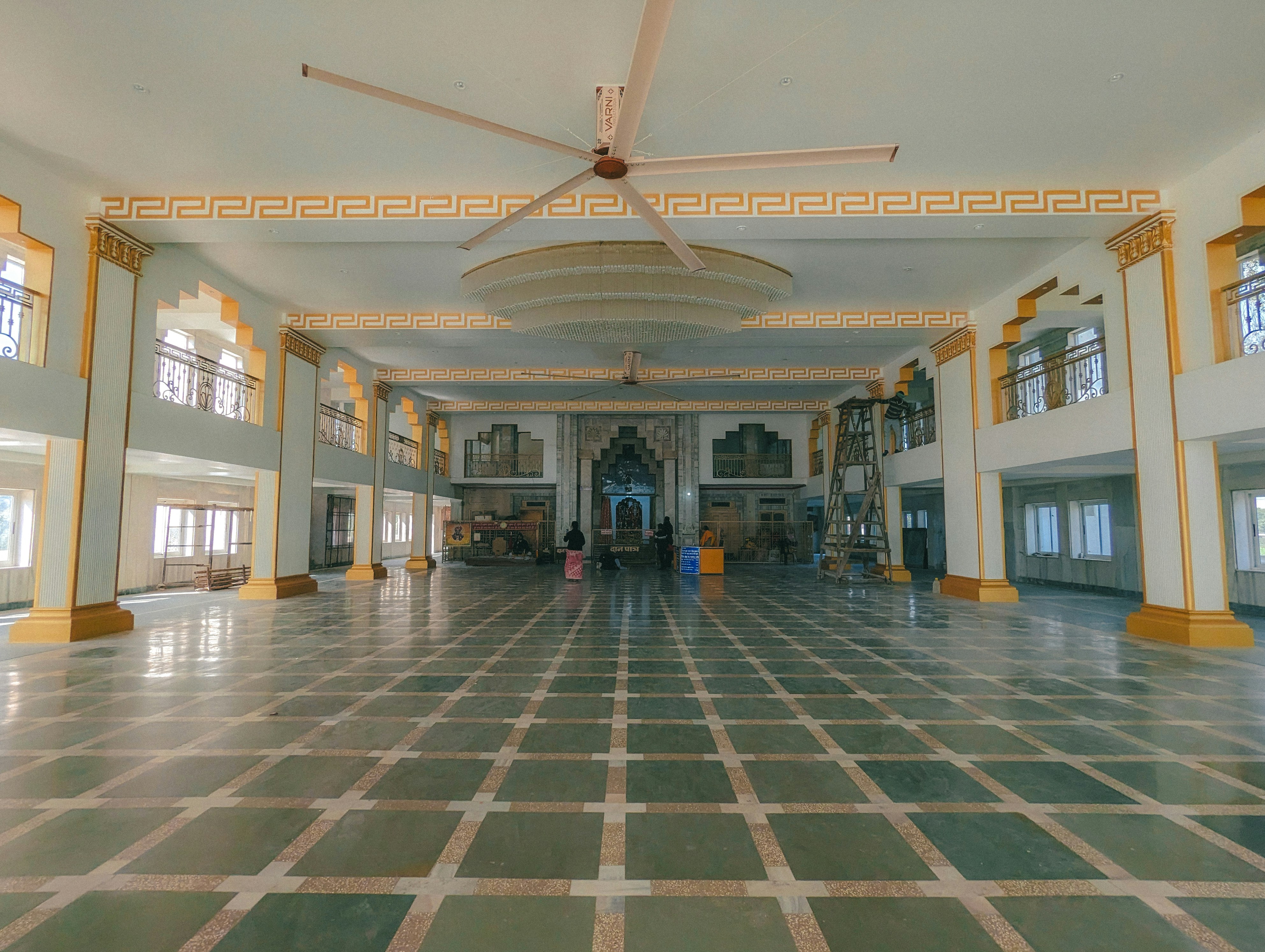 A spacious interior with high ceilings and an ornate design, featuring large green and beige tiled floors, decorated pillars with gold accents, and a massive ceiling fan. The architecture includes intricate molding and a grand entryway at the far end with an ornate arch. Few people are visible, suggesting construction or setup activities, with a ladder and equipment present.