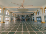 A spacious interior with high ceilings and an ornate design, featuring large green and beige tiled floors, decorated pillars with gold accents, and a massive ceiling fan. The architecture includes intricate molding and a grand entryway at the far end with an ornate arch. Few people are visible, suggesting construction or setup activities, with a ladder and equipment present.