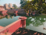 A serene water tank with steps leading down from all sides, constructed with red stone. The tank is surrounded by buildings and trees. The water reflects the sky and nearby greenery, creating a lush and tranquil atmosphere.