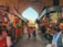 A bustling marketplace scene with stalls on either side displaying colorful textiles, clothing, and bags. Shoppers, dressed in a variety of attire, walk through the corridor under an arched entrance, with a view of an ornate building in the background.
