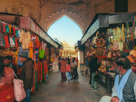 A bustling marketplace scene with stalls on either side displaying colorful textiles, clothing, and bags. Shoppers, dressed in a variety of attire, walk through the corridor under an arched entrance, with a view of an ornate building in the background.