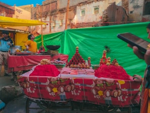 A bustling street market featuring a stall with offerings of vibrant red powders, likely used for festivals or ceremonies. The scene includes piles of colorful powder and various artifacts on display. A man in a yellow shirt stands next to the display, while another person is using a smartphone.