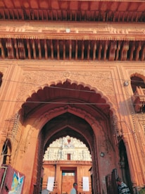 An intricate and ornately carved reddish-brown stone gateway with large arches and decorative details. The structure features detailed carvings and patterns, leading to a smaller, more distant structure with white ornamental designs. People can be seen passing through the gateway.
