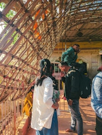 A group of people stand under a bamboo structure with intricate weaving. The environment appears to be a temple or religious site, suggested by the sacred architecture and the presence of an orange prayer cloth tied to the structure. Individuals are engaged in various activities, such as talking and observing their surroundings.