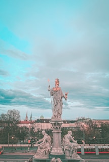 A grand statue stands on a pedestal surrounded by smaller statues. The main statue depicts a robed figure with a staff and an owl, which might suggest wisdom or knowledge. The smaller statues seated around the pedestal also depict robed figures, each holding different objects. In the background, there are buildings with historic architecture and a cloudy sky with a hint of turquoise.