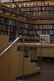 A cozy library setting with wooden bookshelves filled with a variety of books arranged neatly. Soft lighting illuminates the space, casting a warm glow. A wooden staircase with metal railings leads up to more shelves, giving a sense of depth and exploration.