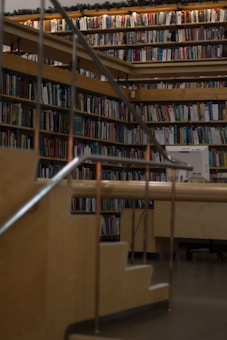 A cozy library setting with wooden bookshelves filled with a variety of books arranged neatly. Soft lighting illuminates the space, casting a warm glow. A wooden staircase with metal railings leads up to more shelves, giving a sense of depth and exploration.