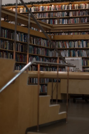 A cozy library setting with wooden bookshelves filled with a variety of books arranged neatly. Soft lighting illuminates the space, casting a warm glow. A wooden staircase with metal railings leads up to more shelves, giving a sense of depth and exploration.