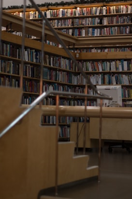 A cozy library setting with wooden bookshelves filled with a variety of books arranged neatly. Soft lighting illuminates the space, casting a warm glow. A wooden staircase with metal railings leads up to more shelves, giving a sense of depth and exploration.