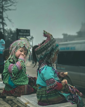 Two children dressed in colorful traditional attire adorned with intricate patterns and beads sit on stone steps. One child is looking towards the camera while eating, and the other is facing away, holding a piece of food. The background includes a foggy environment and a blurred vehicle.
