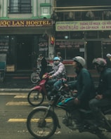 Several people wearing helmets are riding motorbikes along a street in front of a building with signs in Vietnamese. The atmosphere appears busy, with various shops and a hostel visible in the background.