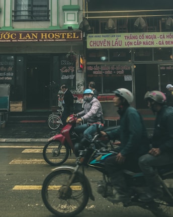 Several people wearing helmets are riding motorbikes along a street in front of a building with signs in Vietnamese. The atmosphere appears busy, with various shops and a hostel visible in the background.