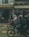 Several people wearing helmets are riding motorbikes along a street in front of a building with signs in Vietnamese. The atmosphere appears busy, with various shops and a hostel visible in the background.