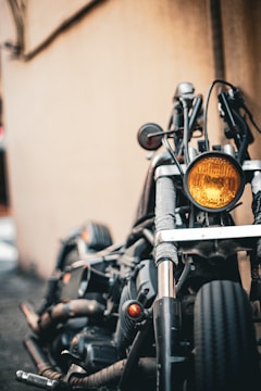 A close-up view of a motorcycle's front, highlighting its headlight, handlebars, and front tire. The background is a blurred wall, creating a rustic urban setting. The image focuses on the mechanical and rugged aspects of the motorcycle.
