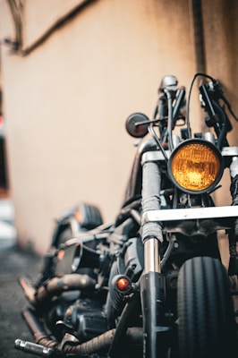A close-up view of a motorcycle's front, highlighting its headlight, handlebars, and front tire. The background is a blurred wall, creating a rustic urban setting. The image focuses on the mechanical and rugged aspects of the motorcycle.