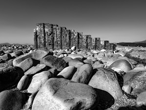 A landscape featuring a large collection of smooth, rounded stones scattered across the ground. A weathered wooden structure, resembling an old breakwater, stands prominently in the middle distance. The backdrop is a clear sky and faint outlines of mountains, creating a monochromatic, serene atmosphere.